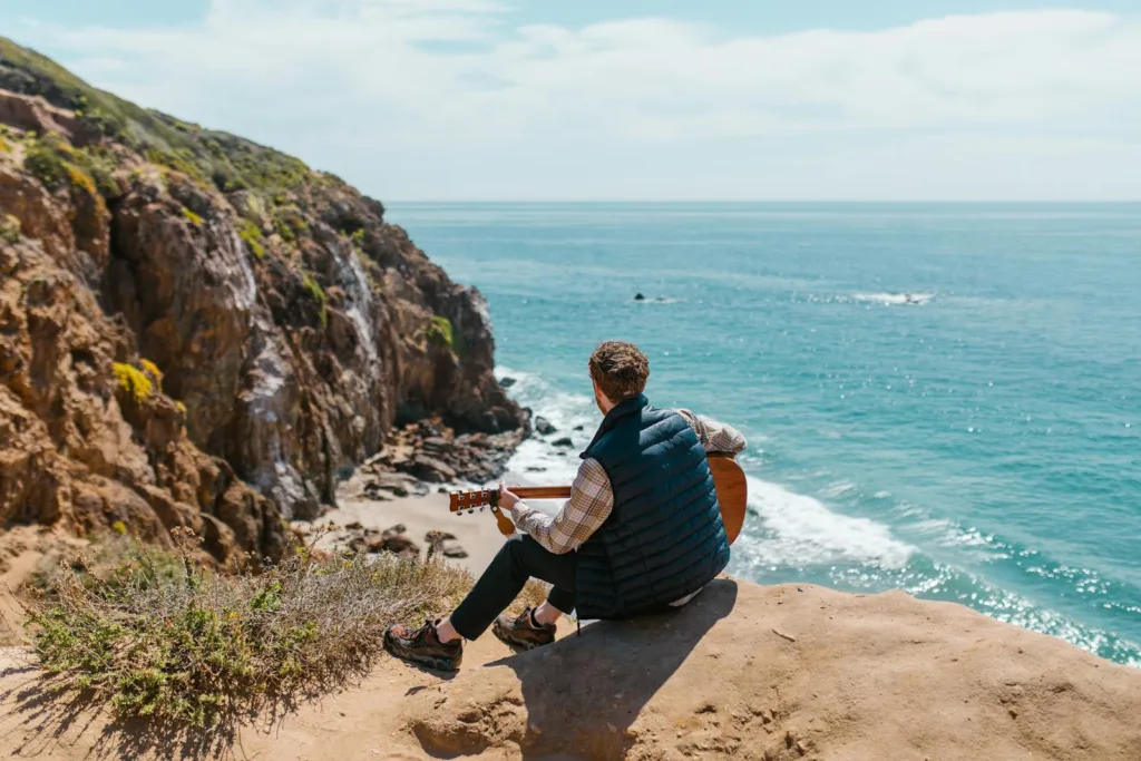 An adult with a guitar sits on a cliff overlooking the ocean, embracing nature and serenity from Gen Z Solo Retreats in California